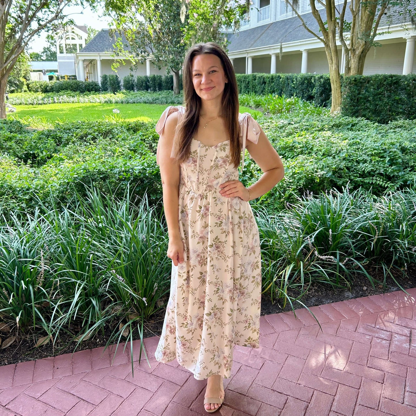 Woman in a floral dress standing on a brick path with greenery and a building in the background