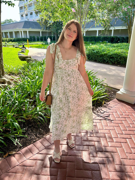 Woman in a floral dress standing on a brick path with greenery and a building in the background