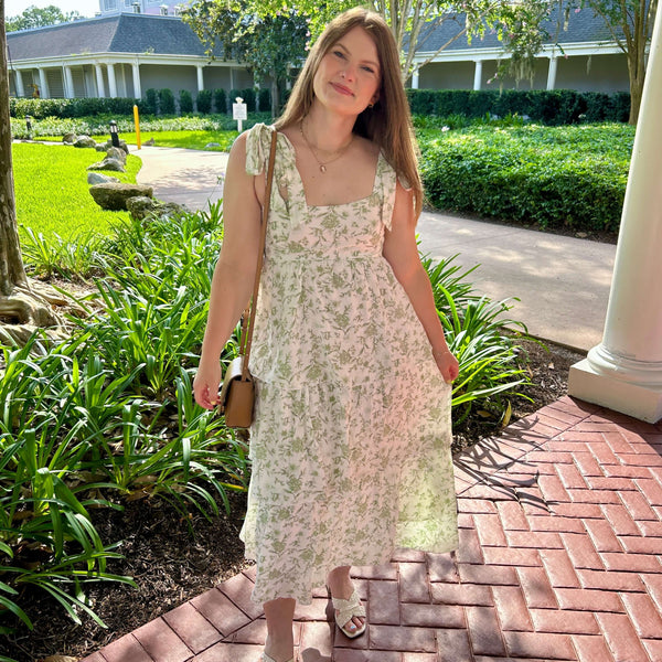 Woman in a floral dress standing on a brick path with greenery and a building in the background