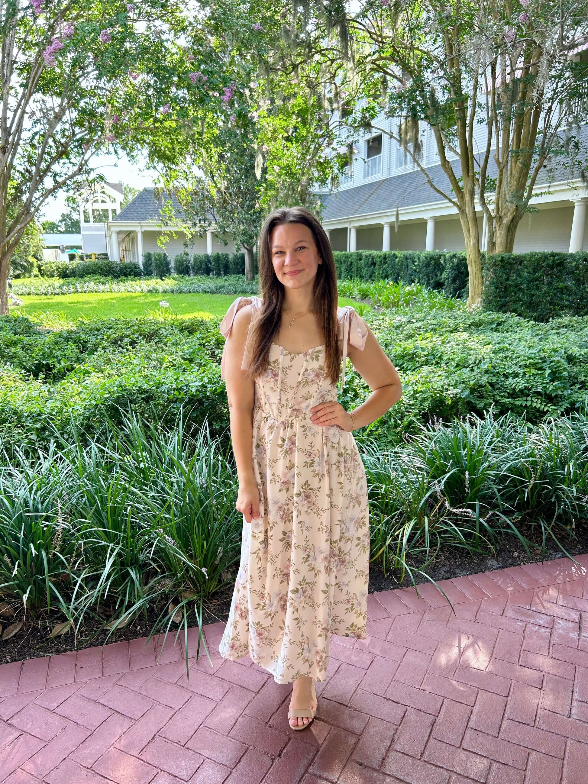 Woman in a floral dress standing on a brick path with greenery and a building in the background