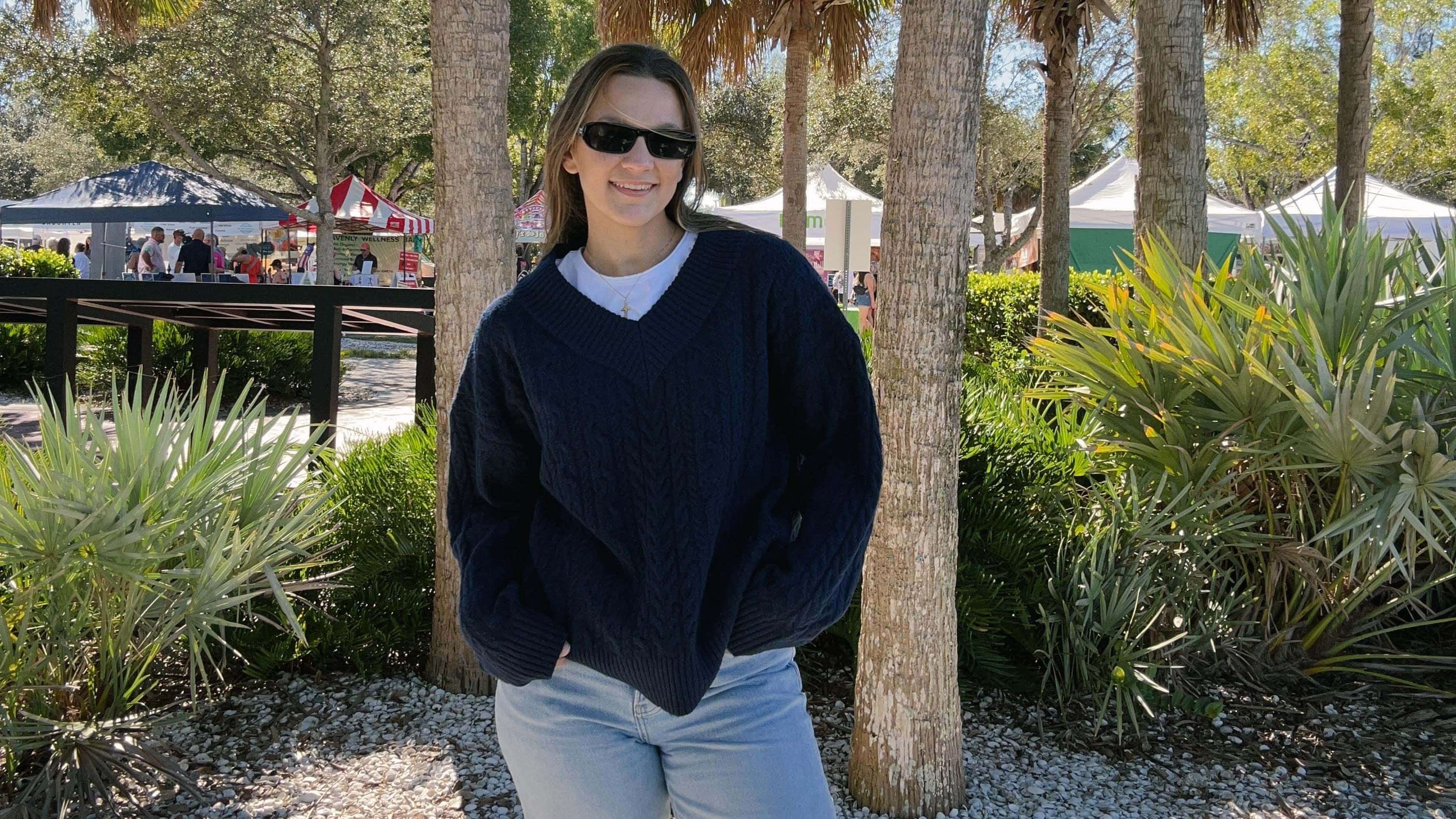 A woman wearing a dark navy blue knit sweater, white t-shirt, and light blue jeans stands outdoors on a gravel path surrounded by palm trees.